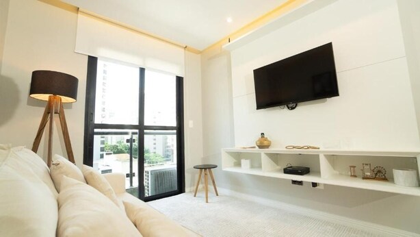 A modern living room with a sofa, a floor lamp, a TV mounted on the wall, and a balcony with a view. - PRIME LOK- SP FLAT JARDINS II (São Paulo)