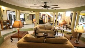 A spacious living area with a large sofa, a coffee table, and a ceiling fan. There are lamps on side tables and a fireplace in the background. - Villa Paradise- Pool, Jacuzzi & More (Montego Bay)