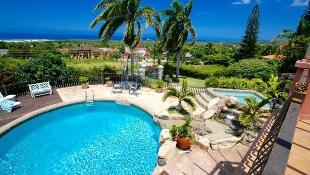 A swimming pool with a curved edge, surrounded by palm trees and a view of the ocean. - Villa Paradise- Pool, Jacuzzi & More (Montego Bay)