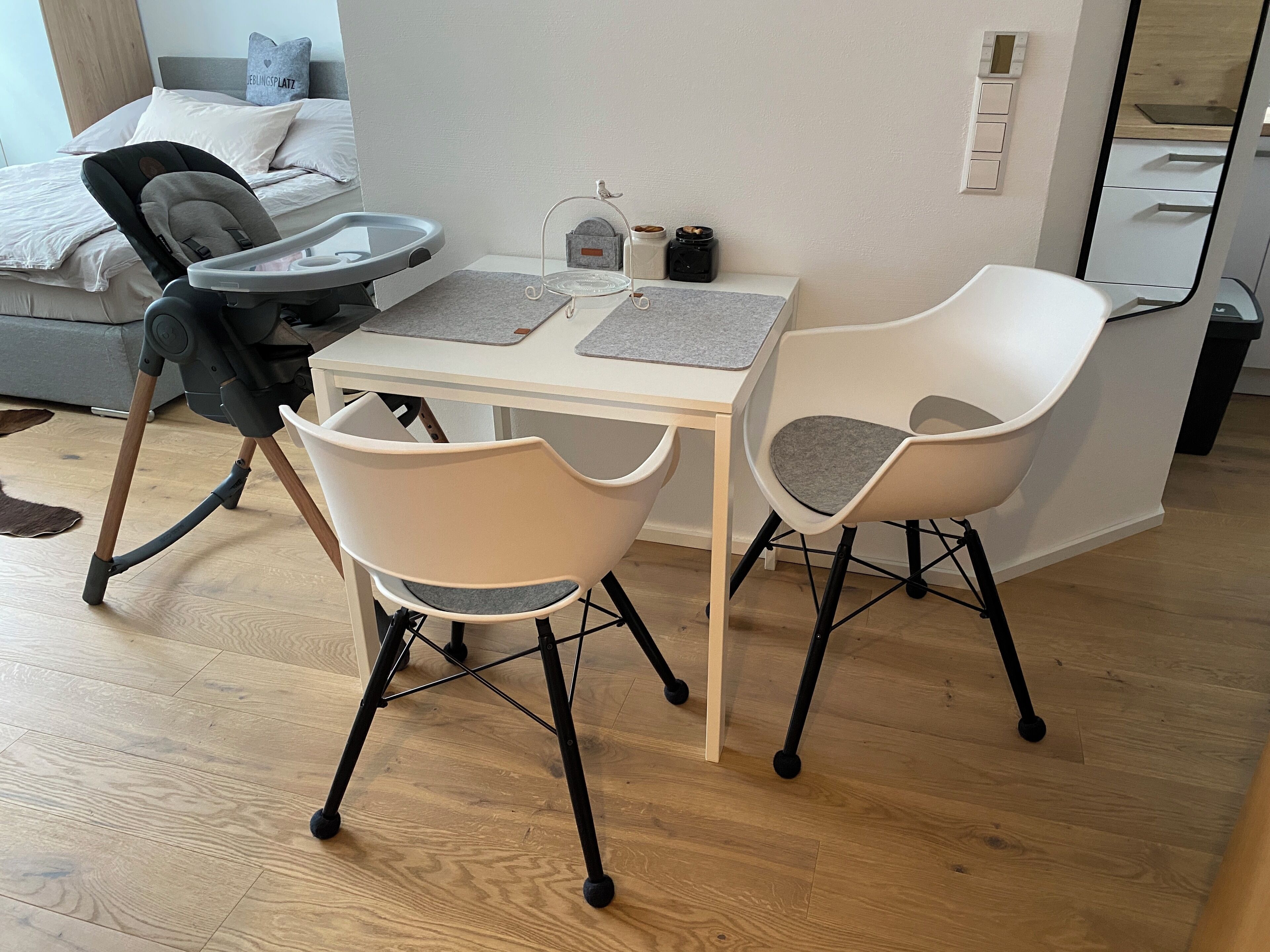 Ein Zimmer mit einem Bett, einem Schreibtisch, zwei Stühlen, einem Babywippstuhl und einem kleinen Tisch mit Gläsern.