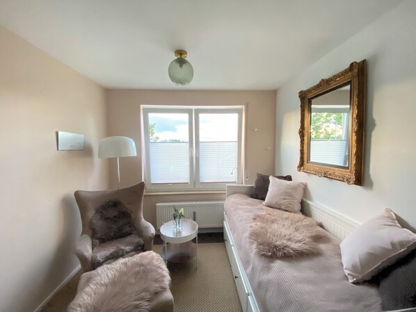A cozy living room with a sofa, armchair, and a small table with a lamp. There is a window with blinds and a ceiling light fixture. - Lieblingsplatz (Aichtal)