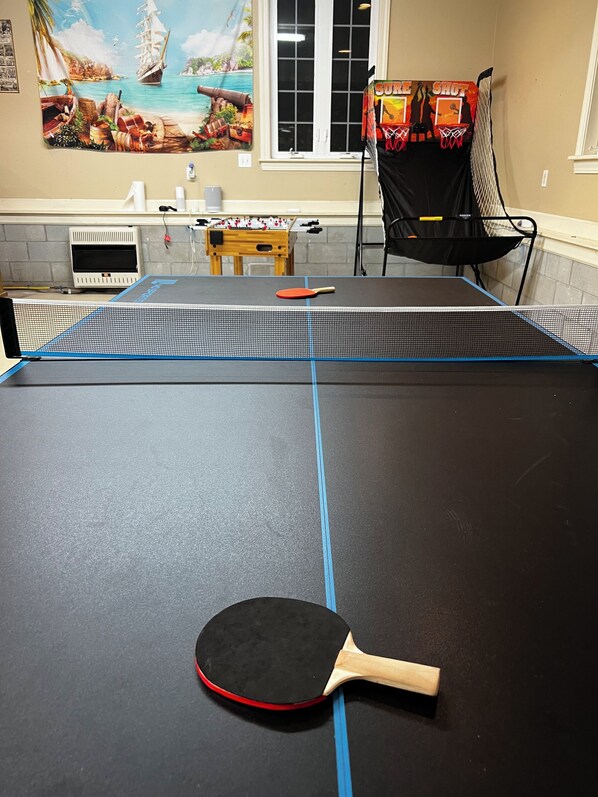 Una mesa de ping pong con una paleta, una mesa de fútbol de mesa y un aro de baloncesto en una habitación con un mural de una escena tropical.