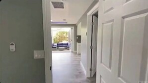 Un couloir avec des portes blanches, un interrupteur et, au loin, on aperçoit un canapé bleu et une fenêtre.
