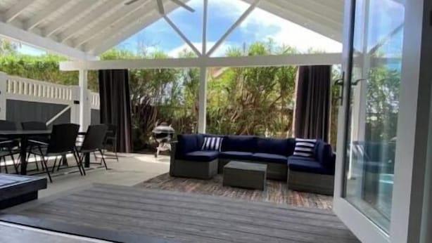 Un espace extérieur abrité, doté d’un ventilateur de plafond, d’une table à manger avec des chaises, d’un canapé et d’une porte vitrée donnant sur un paysage verdoyant.