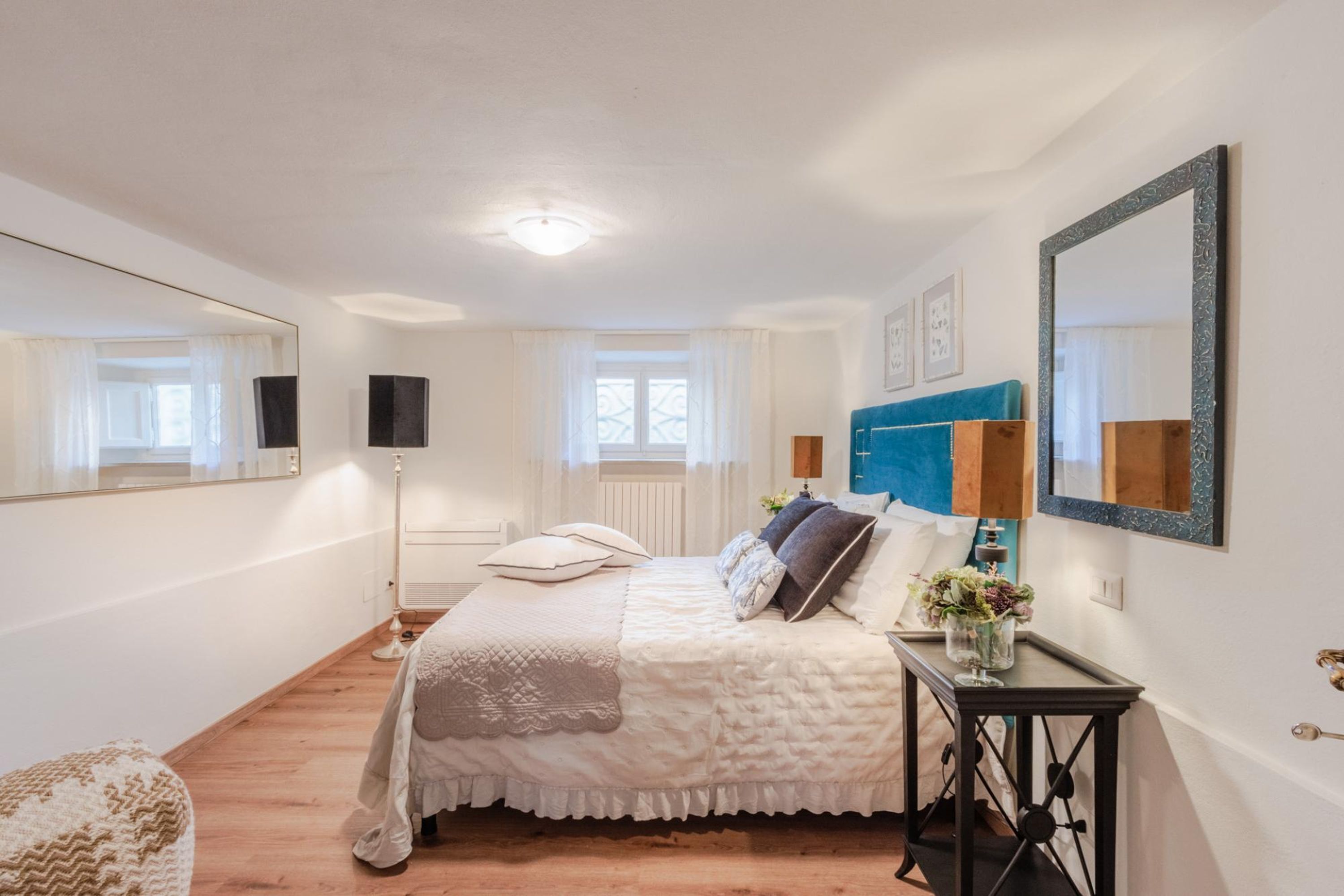 A bedroom with a large bed, a bedside table with a vase of flowers, a floor lamp, and a mirror on the wall.