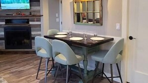 A dining area with a wooden table set for four, surrounded by four chairs. A television is mounted on a wooden wall above the fireplace, and a mirror hangs on the wall above the table. - The Nest!! (Lantzville)