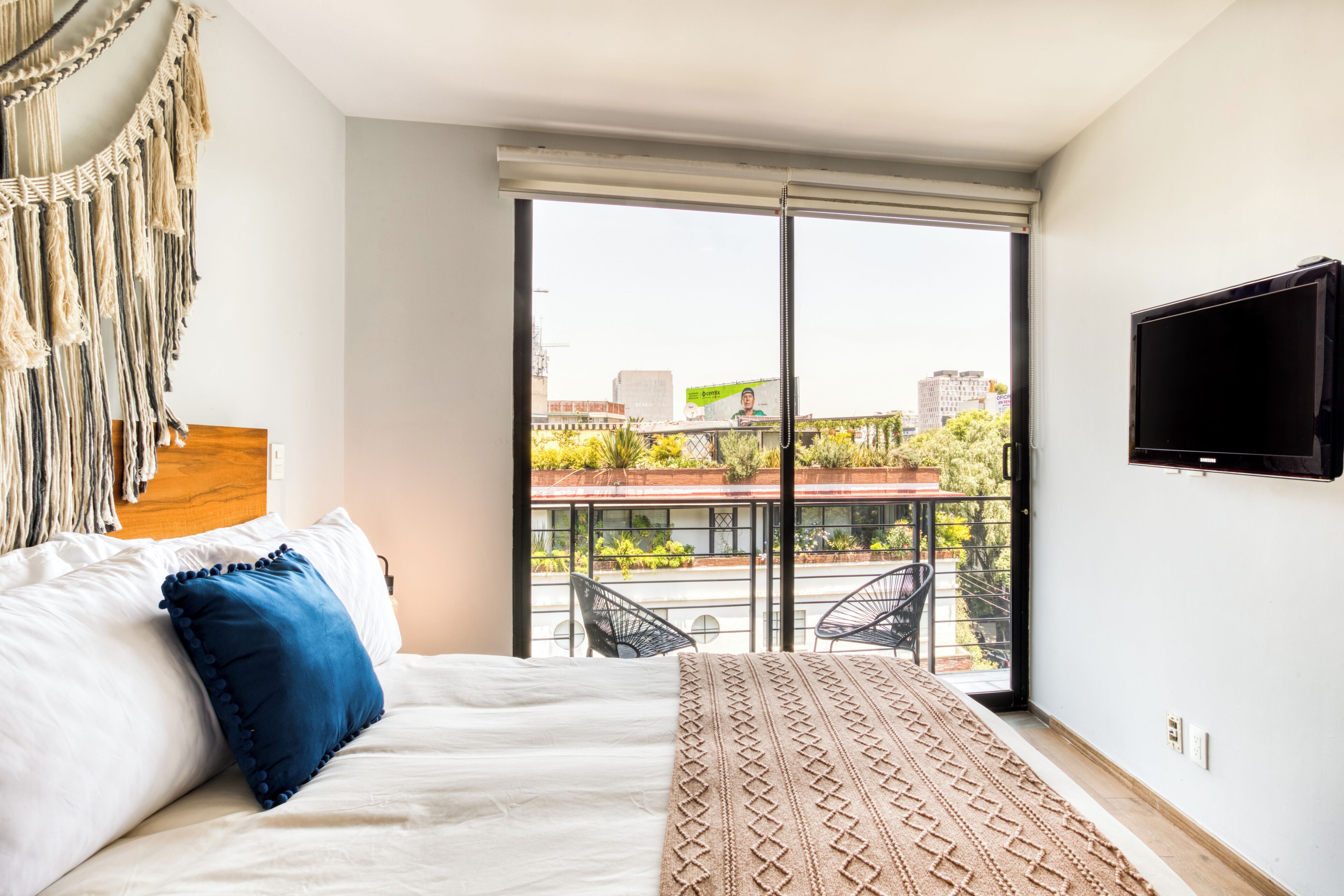 A bedroom with a bed, a large window, a TV, and a macrame wall hanging.