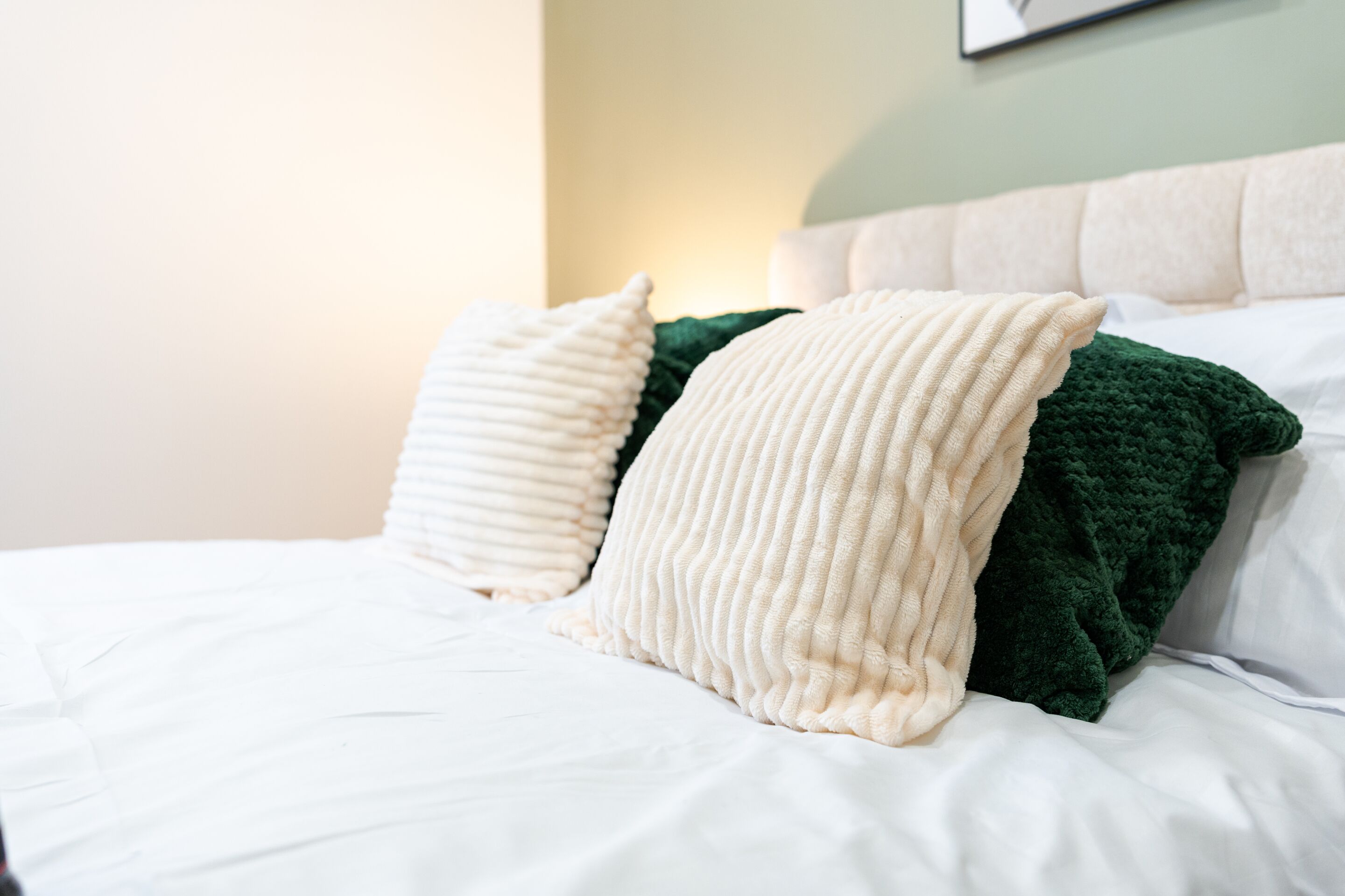 Een bed met witte lakens en twee kussens, één crème en één groen, in een kamer met een lichtgroene muur en een wit hoofdeinde.