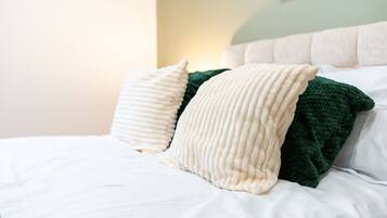 Een bed met witte lakens en twee kussens, één crème en één groen, in een kamer met een lichtgroene muur en een wit hoofdeinde.