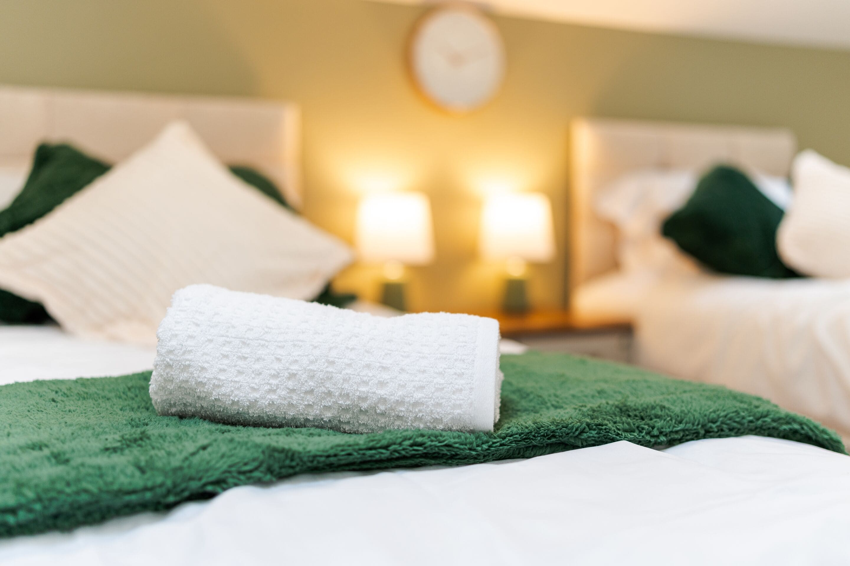 A neatly made bed with a green blanket and a rolled white towel, two bedside lamps, and a clock on the wall.