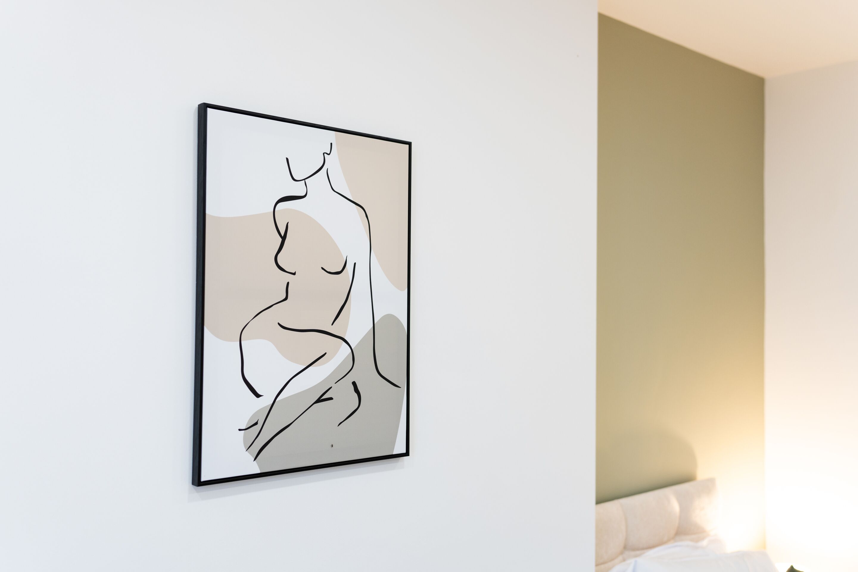 A framed abstract art piece on a white wall in a room with a bed and a green wall.