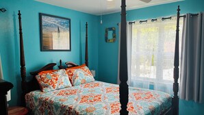 Een hemelbed met een bed met een patroon erop, een plafondventilator, een raam met gordijnen en een ingelijst schilderij aan de muur.