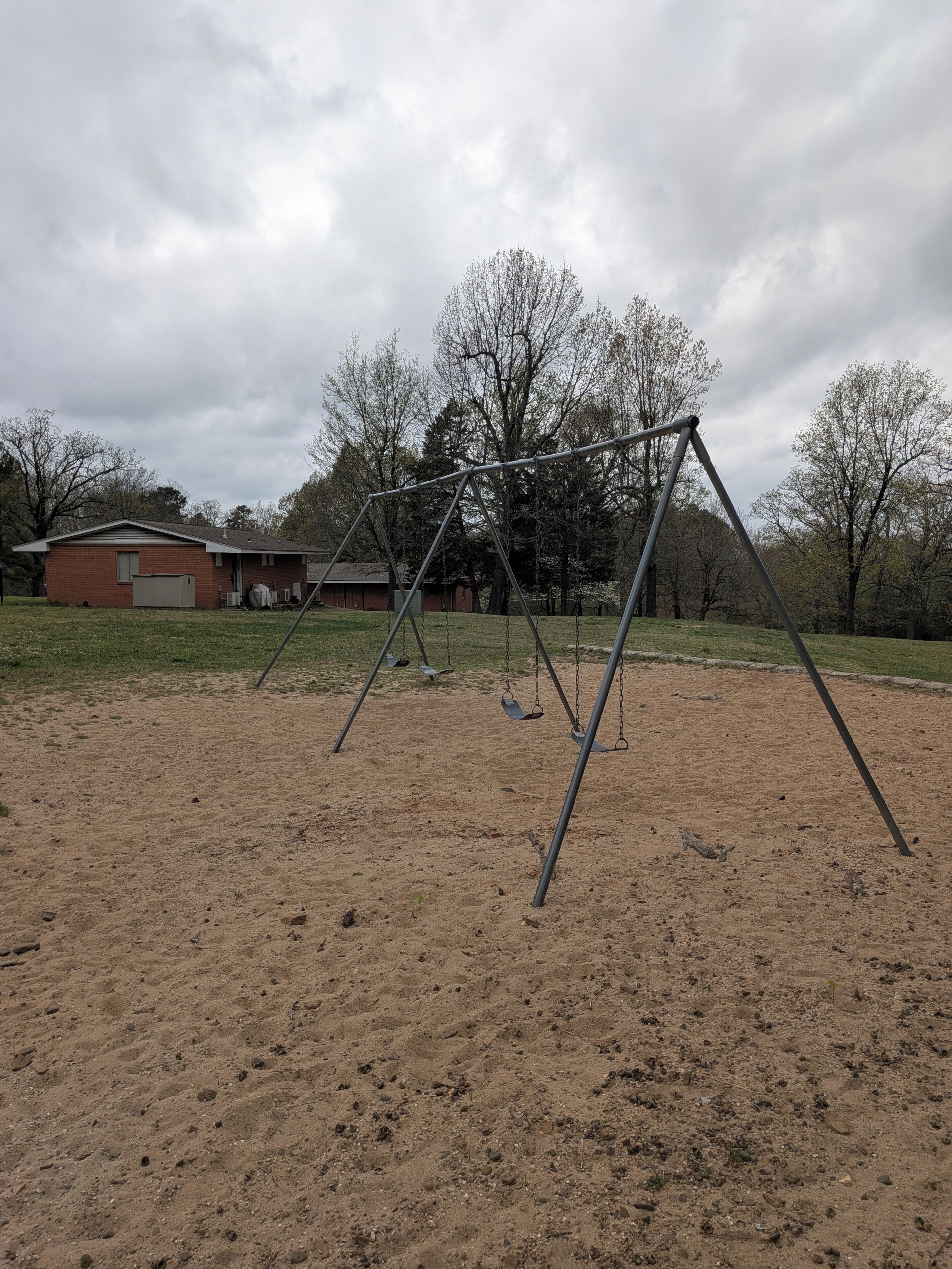 A swing set in a sandy area with a house and trees in the background.