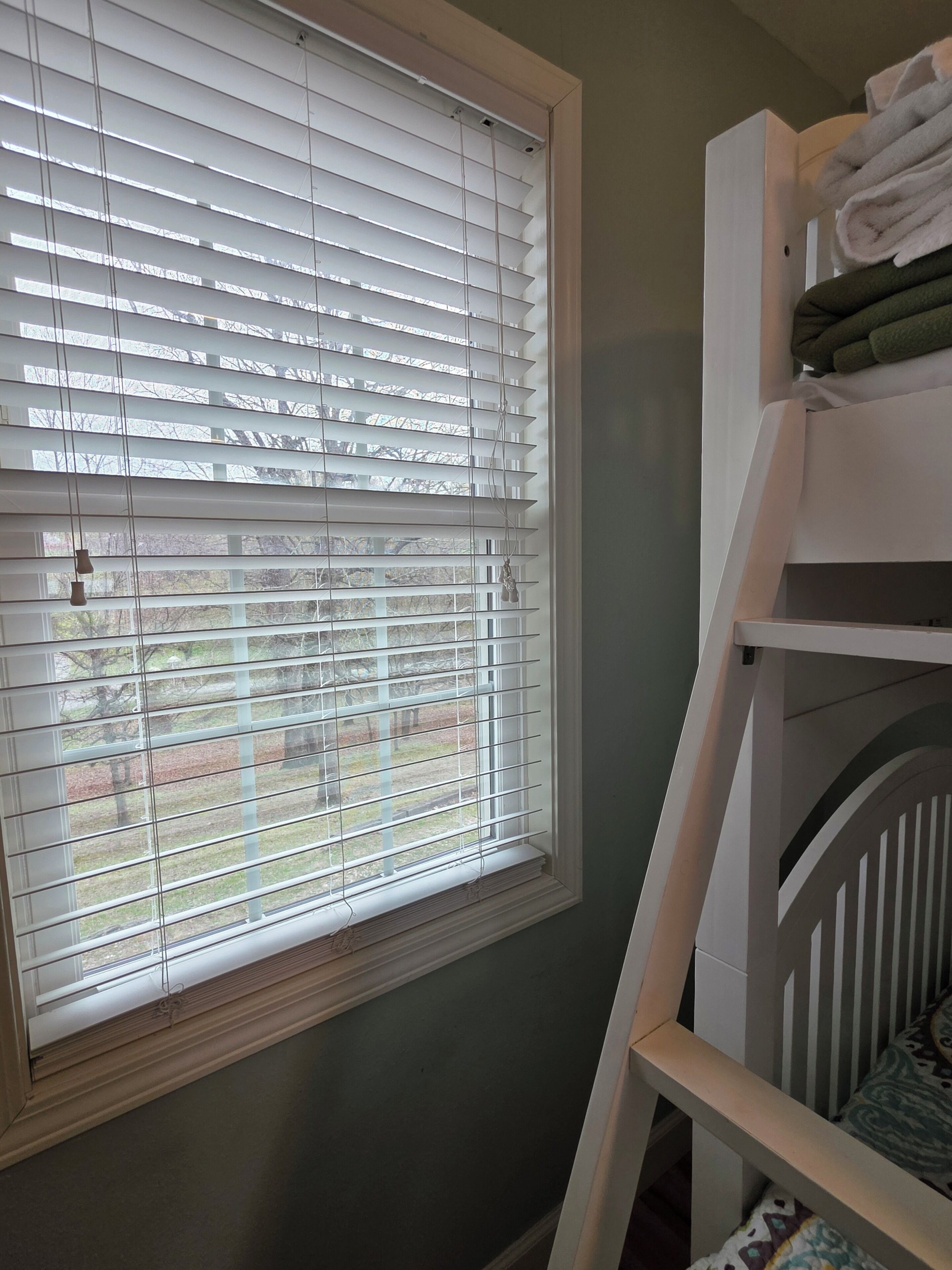 A room with a window with blinds, a crib, and a shelf with folded towels.