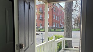 A door leading to a porch with a white railing, a doormat, and a view of a brick building and tree. - Mount Sequoyah Center (Fayetteville)