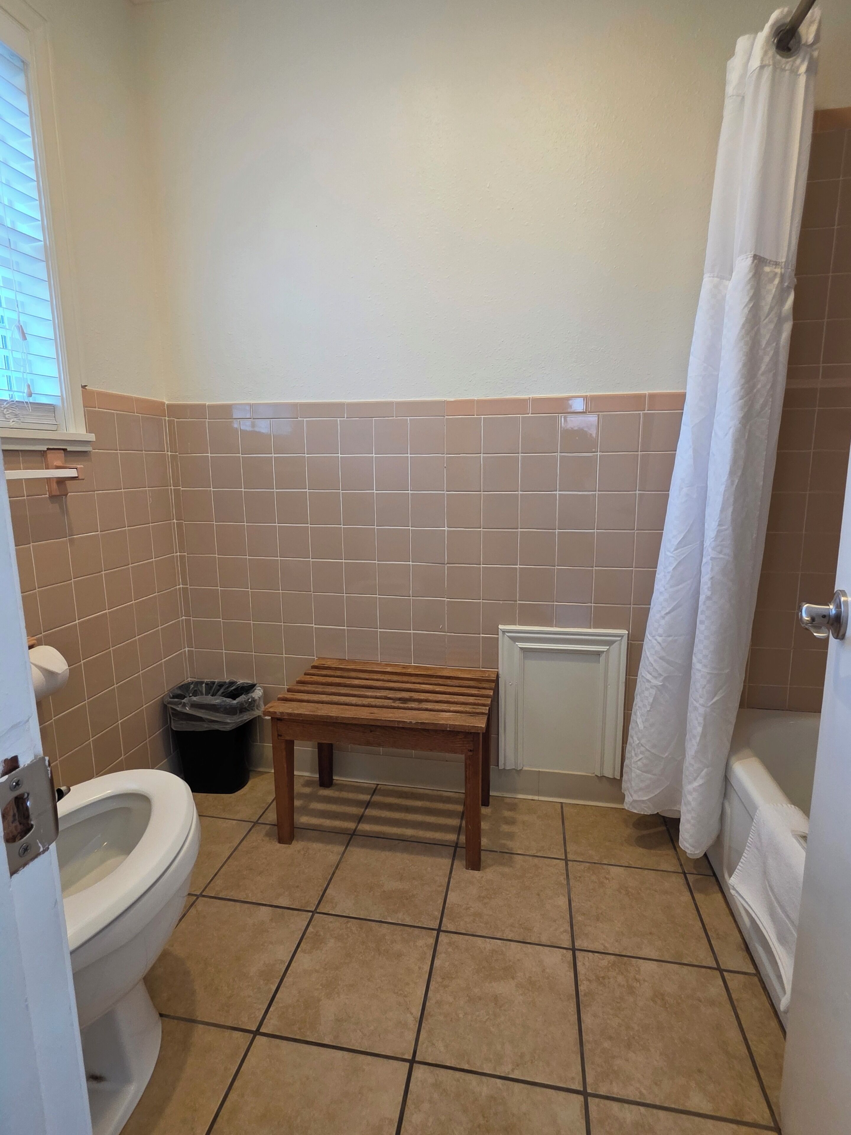 Un baño con inodoro, un banco de madera, un cubo de basura y una cortina de ducha.