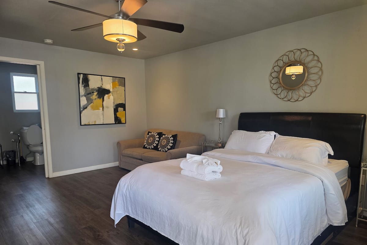 A bedroom with a bed, a ceiling fan, a wall-mounted lamp, a painting, and a door leading to another room.