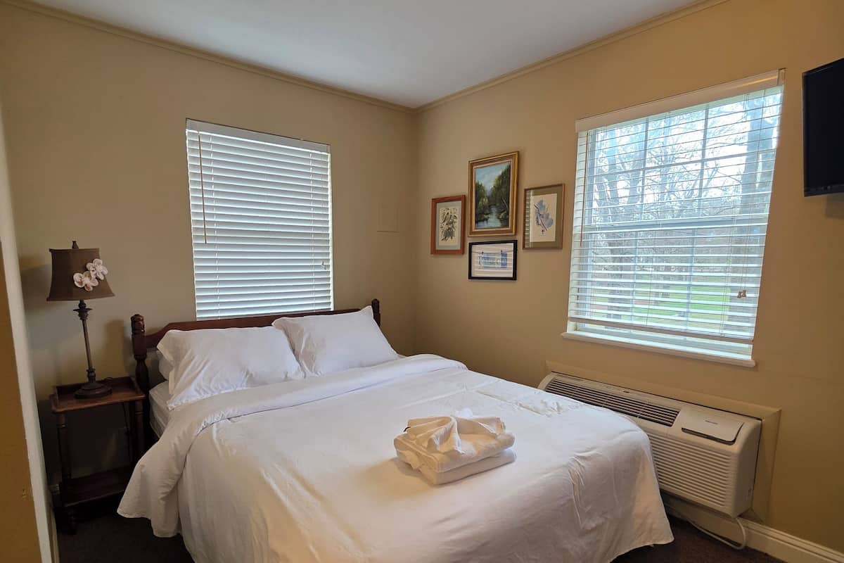 A neatly made bed in a room with two windows, a heater, and wall-mounted artwork.