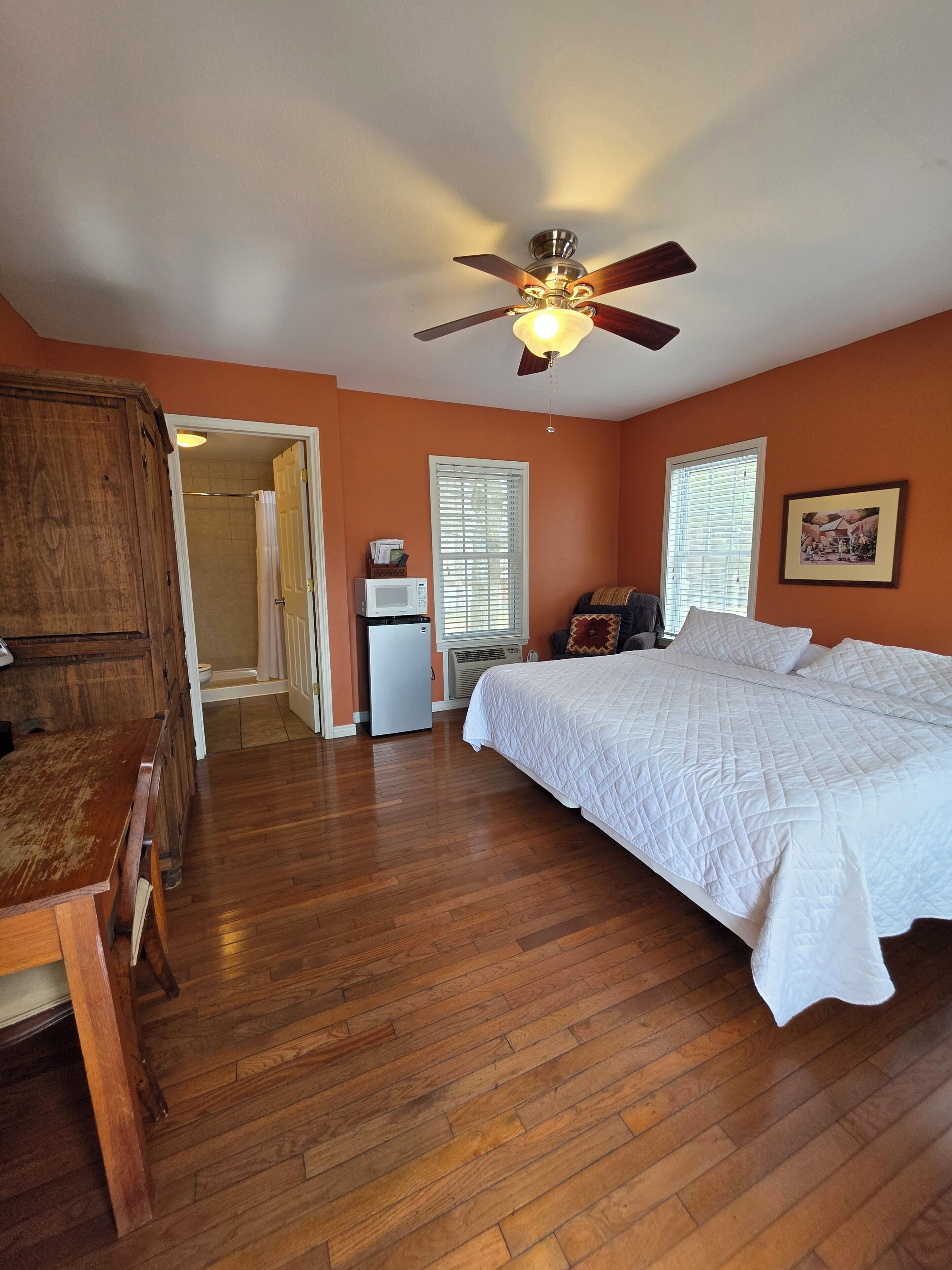 A bedroom with a bed, wooden floor, ceiling fan, and a door leading to a bathroom.