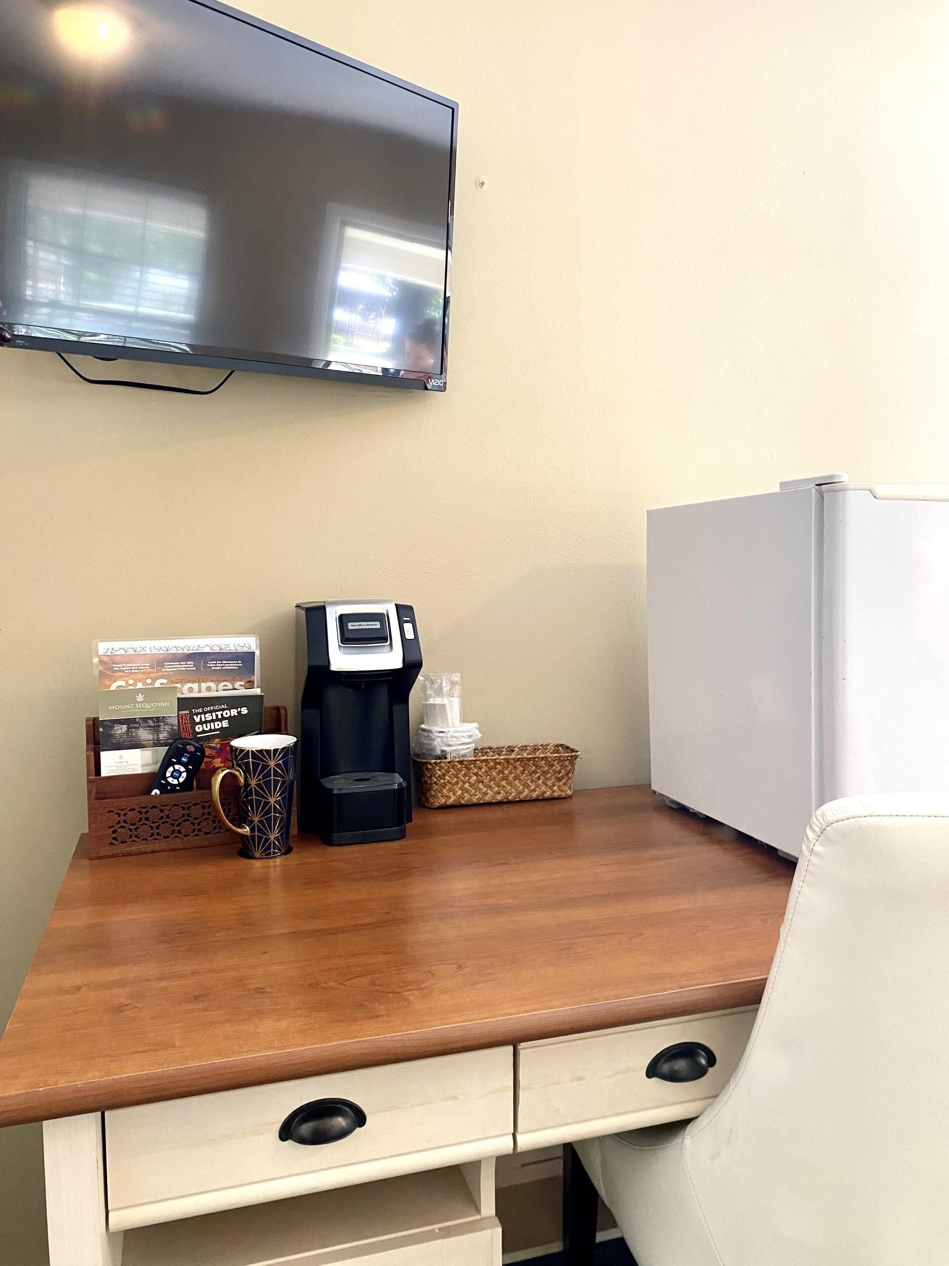 Una habitación de hotel con un escritorio de madera, una cafetera, un control remoto, una taza, una cesta y un televisor montado en la pared.