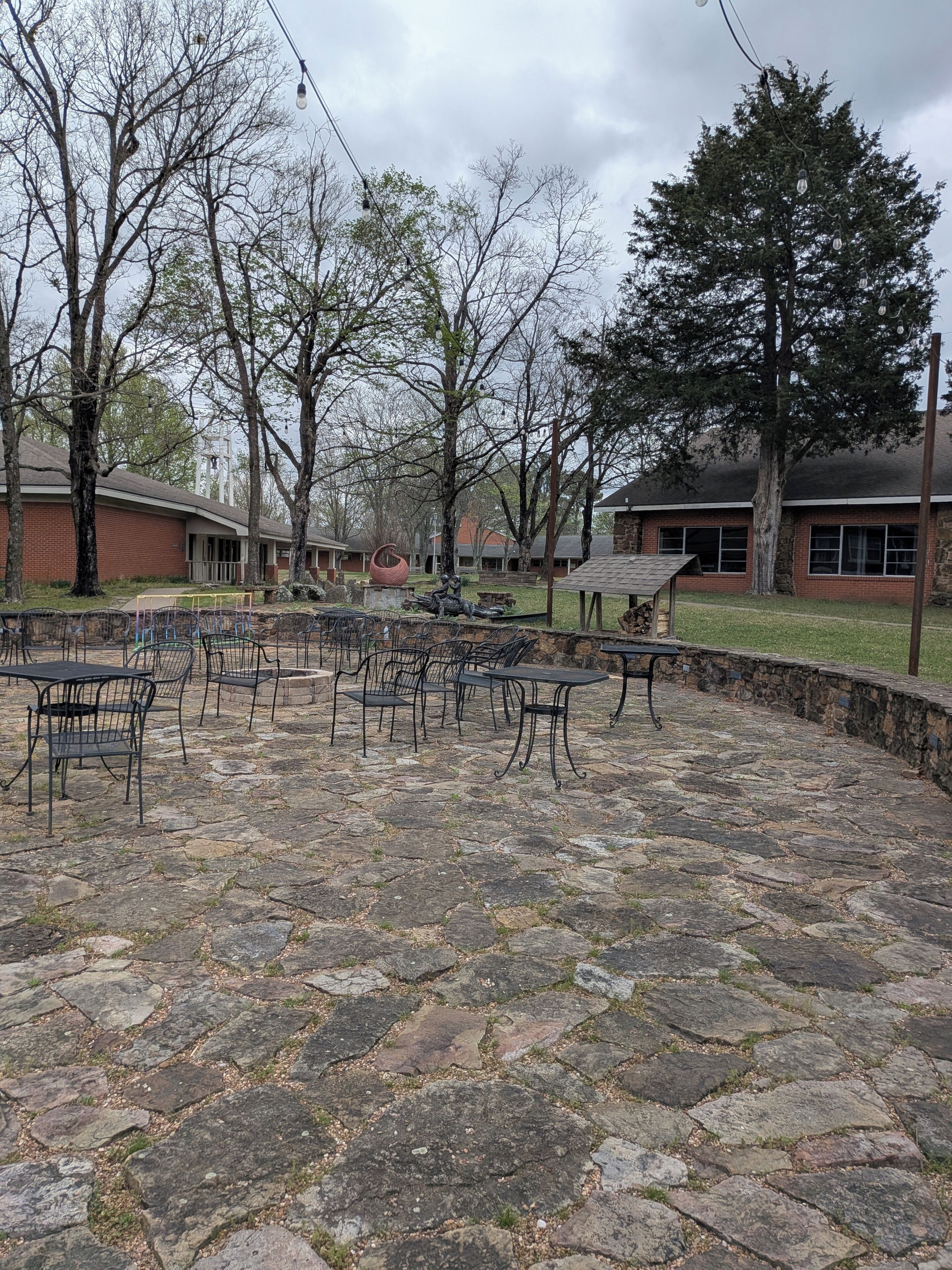 A courtyard with stone flooring, metal tables and chairs, and trees.