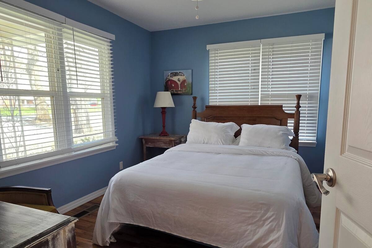 A bedroom with a bed, a ceiling fan, a nightstand with a lamp, and a window with blinds.