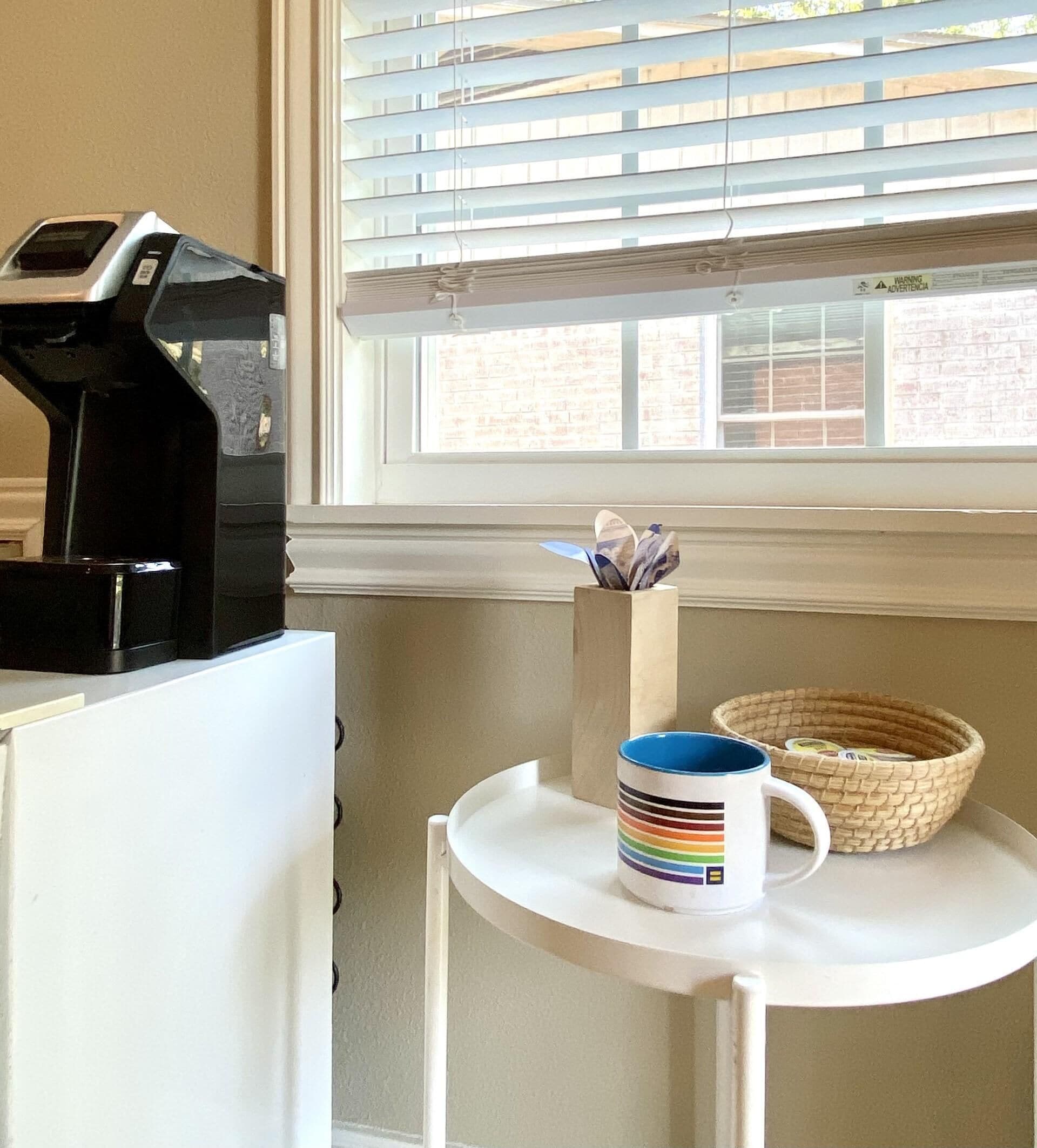 A kitchen area with a coffee maker, a mug on a small table, and a window with blinds.