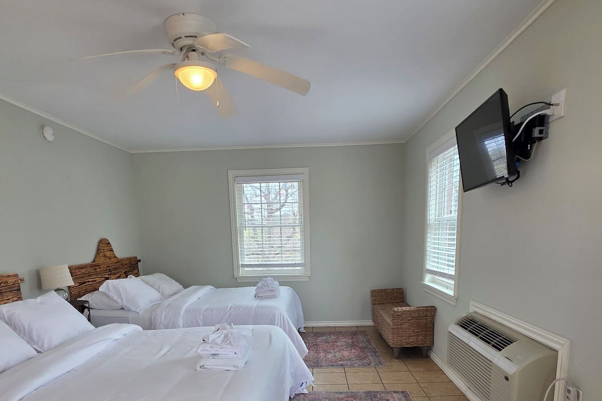 A hotel room with two beds, a ceiling fan, a TV mounted on the wall, and a window with blinds.