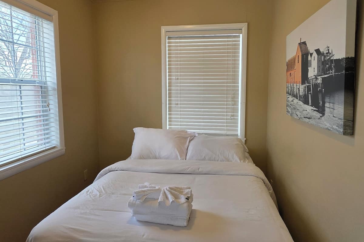 A neatly made bed with white linens, a window with blinds, a ceiling light fixture, and a framed picture on the wall.