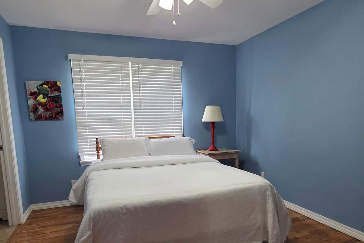 A bedroom with a bed, a ceiling fan, a window with blinds, a nightstand with a lamp, and a wooden floor.