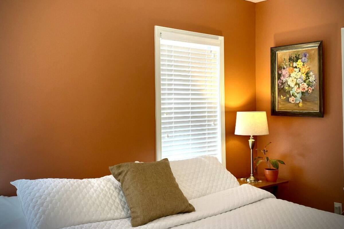 A bedroom with a bed, a window with blinds, a bedside lamp, and a framed floral painting on the wall.