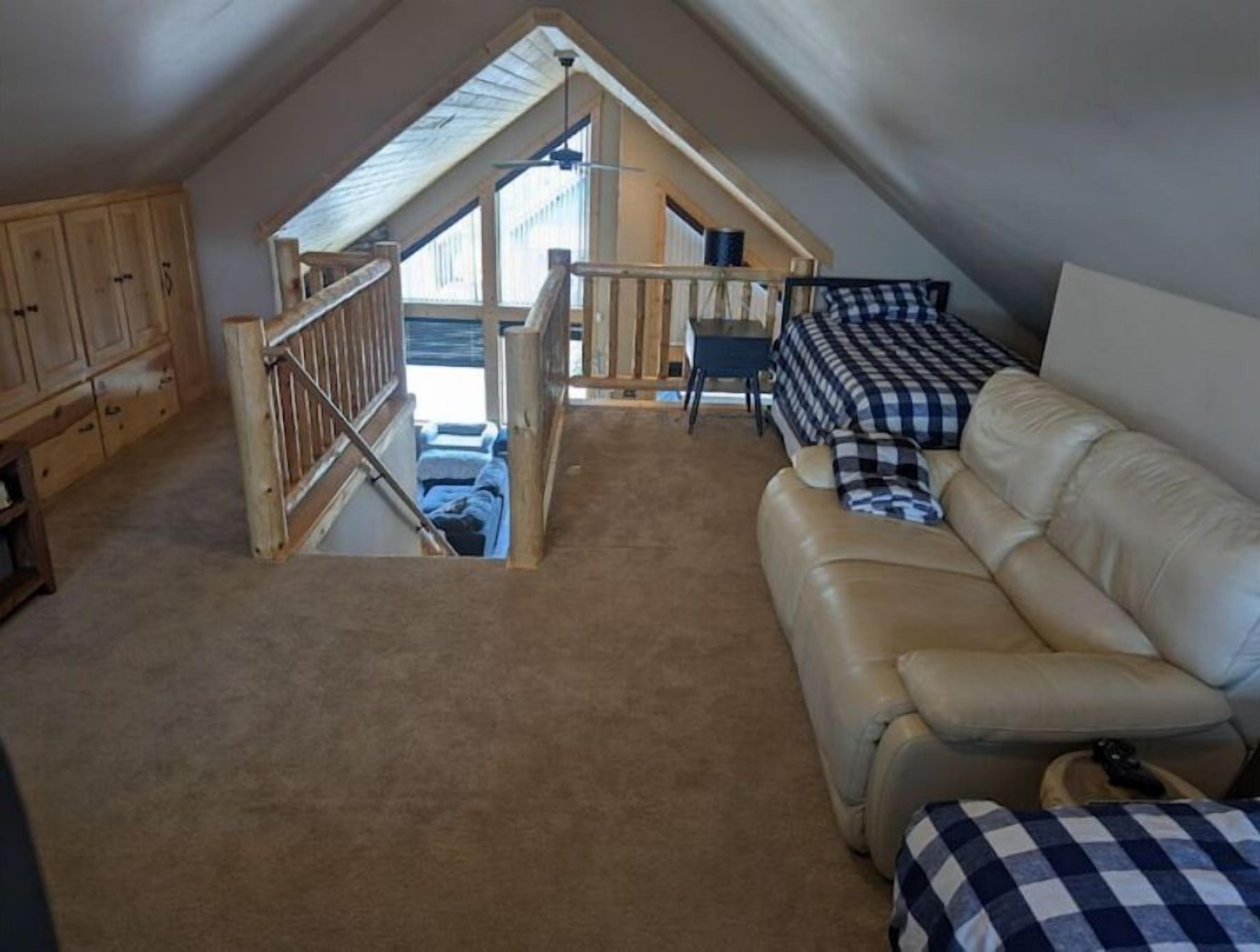 A cozy attic bedroom with a sloped ceiling, a wooden staircase, a leather sofa, and a checkered bedspread.