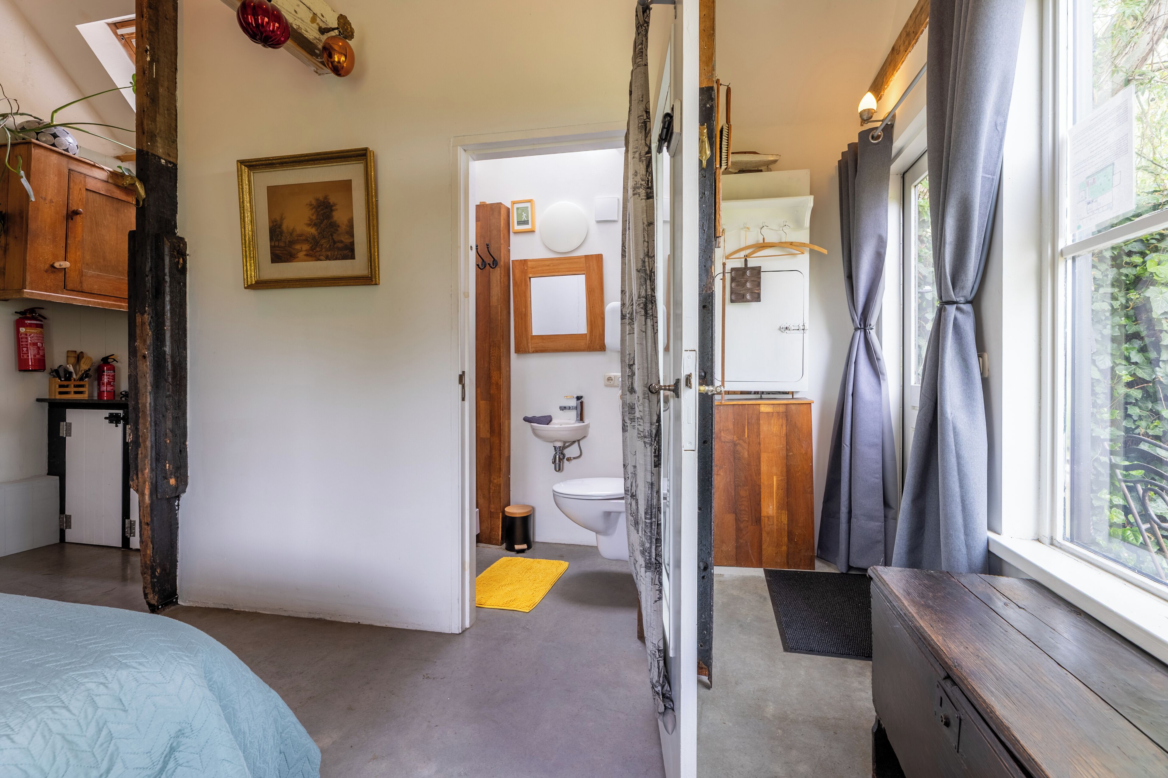 Habitació amb llit, un aparador de fusta, finestra amb cortines, bany amb vàter i rentapans, i una catifes groga.