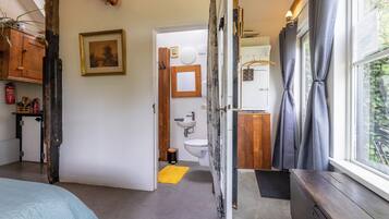 Habitació amb llit, un aparador de fusta, finestra amb cortines, bany amb vàter i rentapans, i una catifes groga.