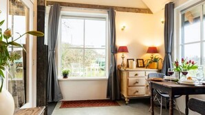 Un coin repas chaleureux avec une table dressée pour deux personnes, une fenêtre avec des rideaux et un petit meuble sur lequel est posée une lampe.