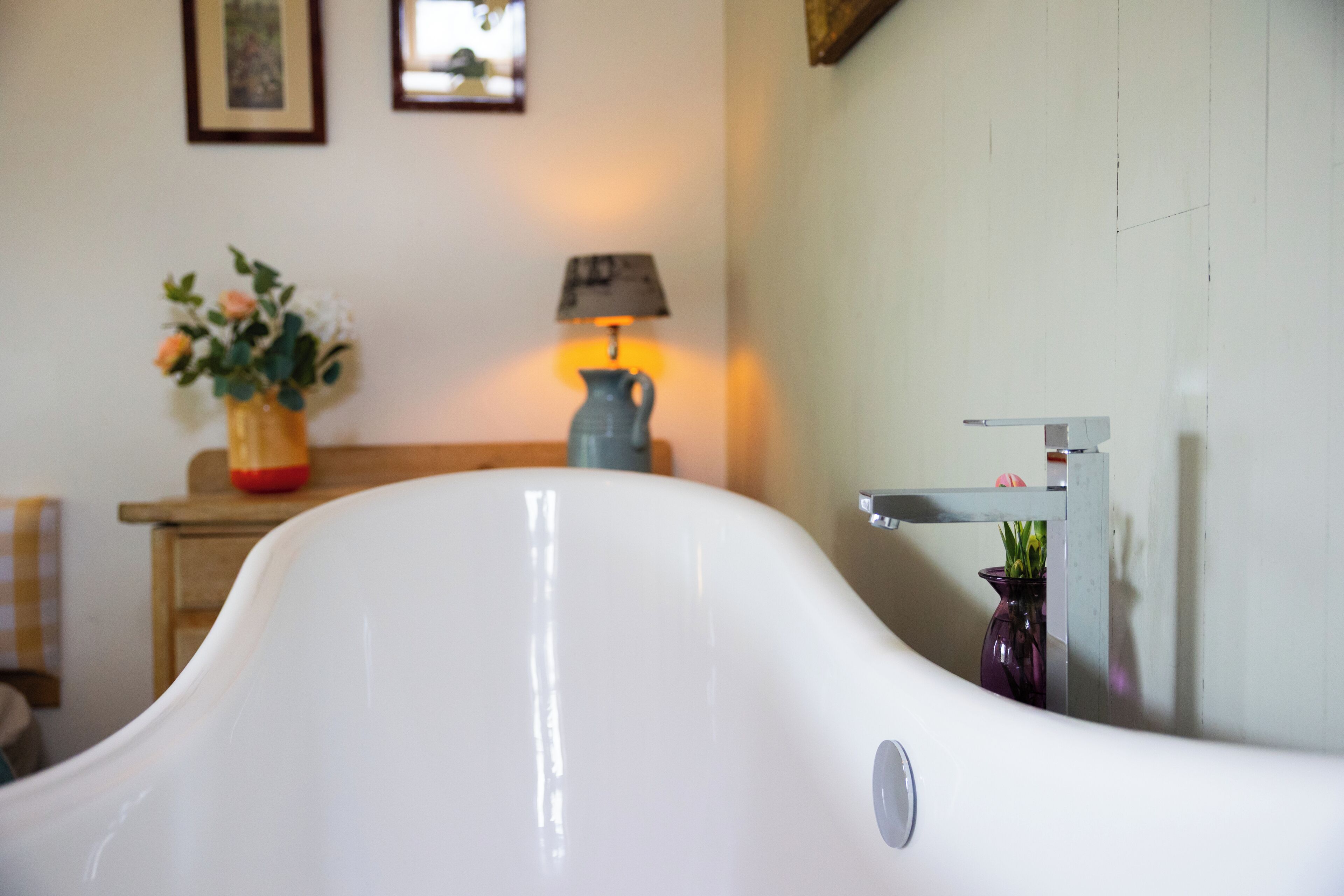 Una bañera blanca en un baño con un tocador de madera, un florero con flores y una lámpara.