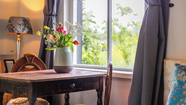 Une pièce avec une table en bois, un vase de fleurs, une lampe et une vue par la fenêtre.