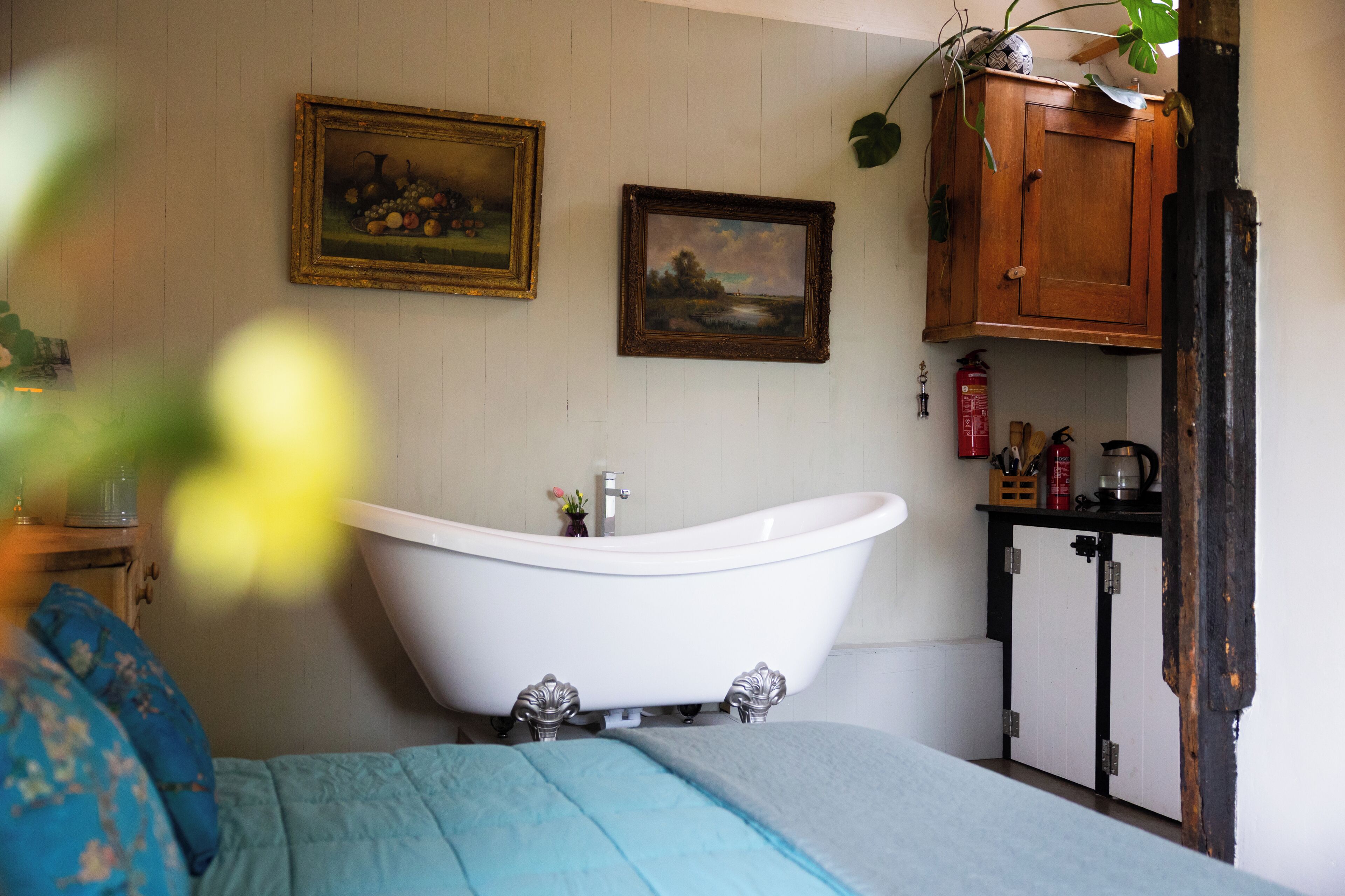 Ein Zimmer mit einer weißen Badewanne mit Füßen, einem Bett mit blauer Bettwäsche, zwei gerahmten Bildern an der Wand und einer Holzkommode mit einer Pflanze darauf.