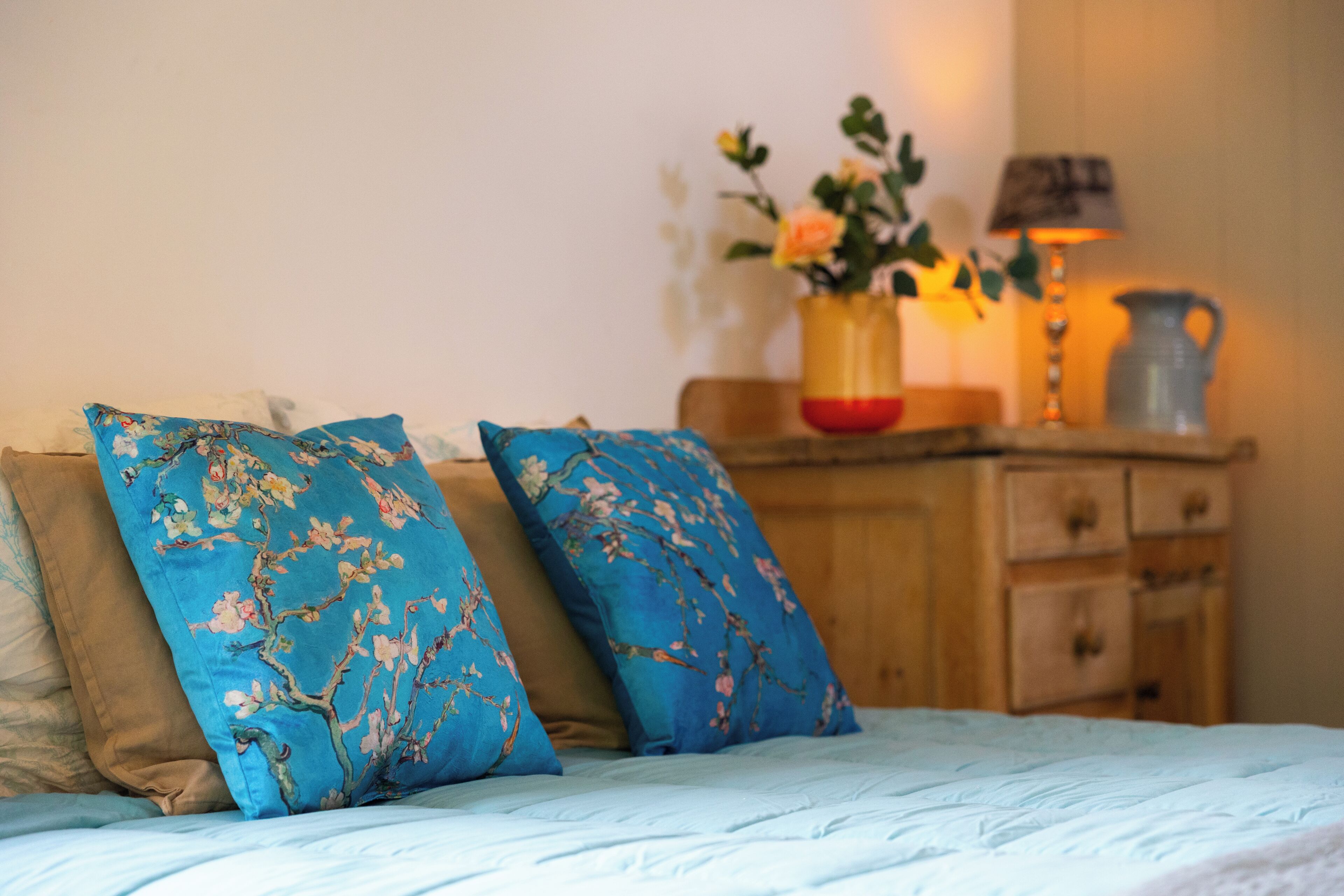 Un lit avec des coussins à motifs floraux, une table de chevet en bois sur laquelle est posé un vase rempli de fleurs, et une lampe.