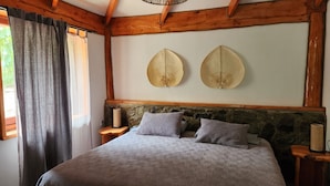 Une chambre avec un grand lit, des poutres en bois, un mur en pierre, deux chapeaux accrochés au mur et une fenêtre avec des rideaux.