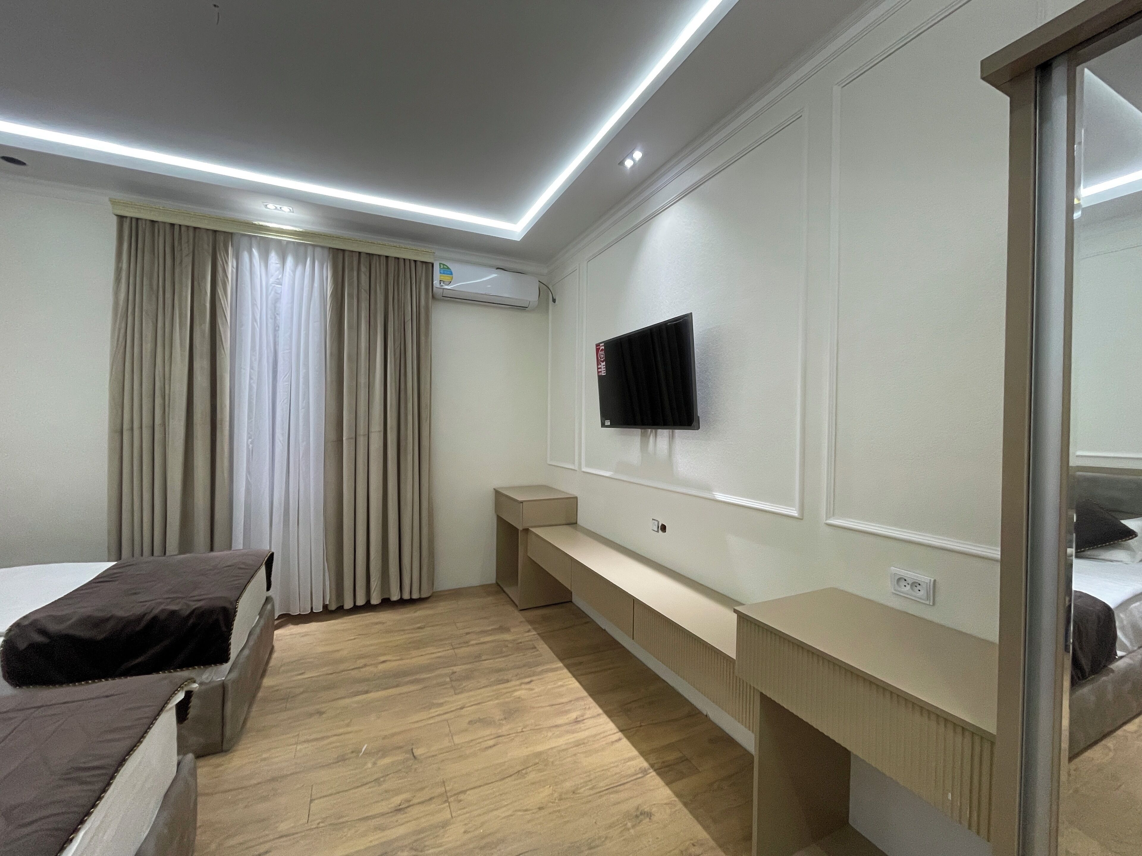 Una habitación de hotel moderna con suelo de madera, una cama con cubierta oscura, una mesita de noche, un televisor montado en la pared y una ventana con cortinas.