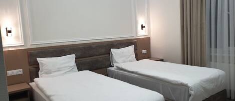 Una habitación de hotel con dos camas individuales, una mesita de noche con lámpara y una ventana con cortinas.