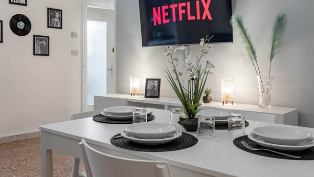A modern dining area with a white table set for four, a flat-screen TV mounted above displaying "NETFLIX", and a wall decorated with framed photos and a vinyl record. - Ariston Centro (Sanremo)