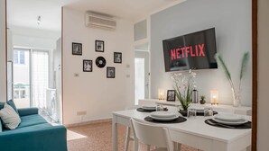 A modern living room with a dining area, a flat-screen TV mounted on the wall, and a balcony with a washing machine visible through the glass door. - Ariston Centro (Sanremo)