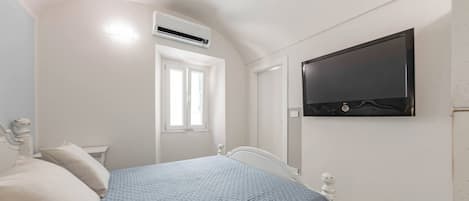 A bedroom with a bed, a window, an air conditioning unit, and a flat-screen TV mounted on the wall.