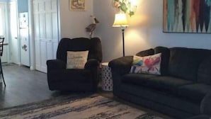 Ein Wohnzimmer mit Deckenventilator, einer schwarzen Couch, einem schwarzen Sessel, einem gemusterten Teppich und einem Gemälde an der Wand.
