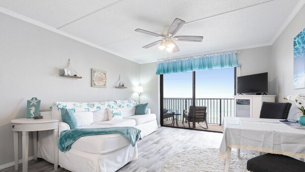 A bedroom with a white bed, a ceiling fan, a TV, and a balcony with a view of the ocean. - Ocean Watch Oasis Monthly Beach Front Retreat (Ormond Beach)