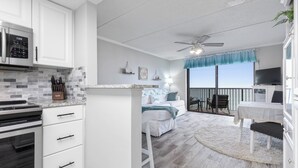 A modern kitchen with white cabinets, a built-in oven, and a microwave. There is a dining area with a table and chairs, and a balcony with a view. - Ocean Watch Oasis Monthly Beach Front Retreat (Ormond Beach)