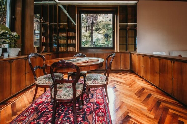 A room with a round wooden table, three chairs, a patterned rug, wooden flooring, a bookshelf, and a window with a view of greenery.
