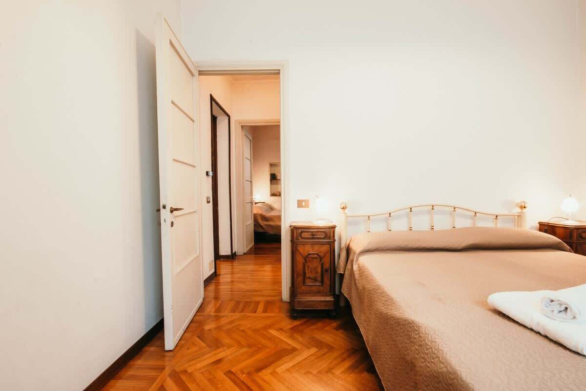 Een slaapkamer met een houten vloer, een bed met hoofdeinde, nachtkastjes met lampen, en een deur naar een andere kamer.